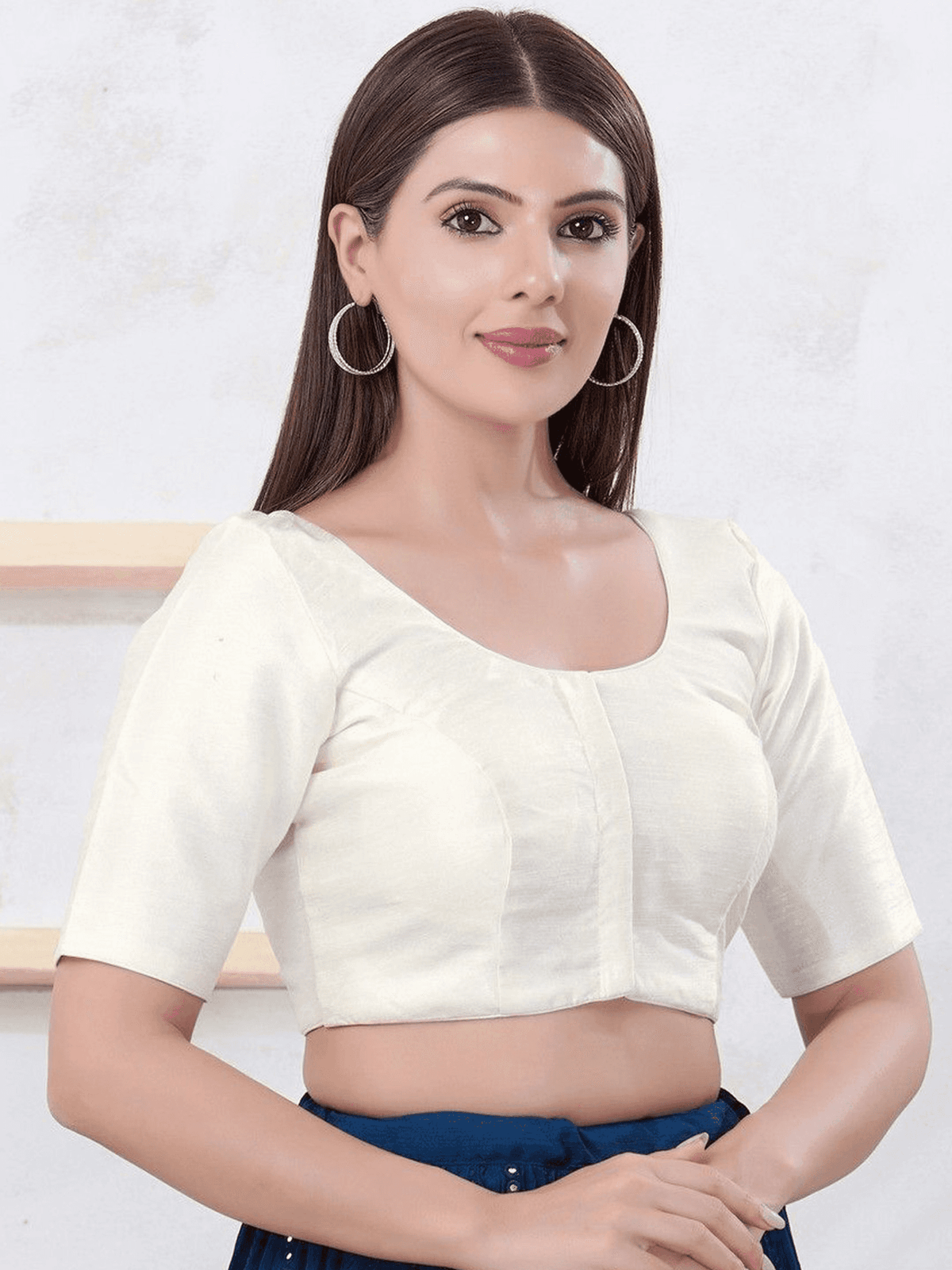 A woman wearing a cream-colored short-sleeve crop blouse with a scoop neckline and a central vertical seam, paired with dark blue bottoms. She is wearing large silver hoop earrings against a light textured wall background.