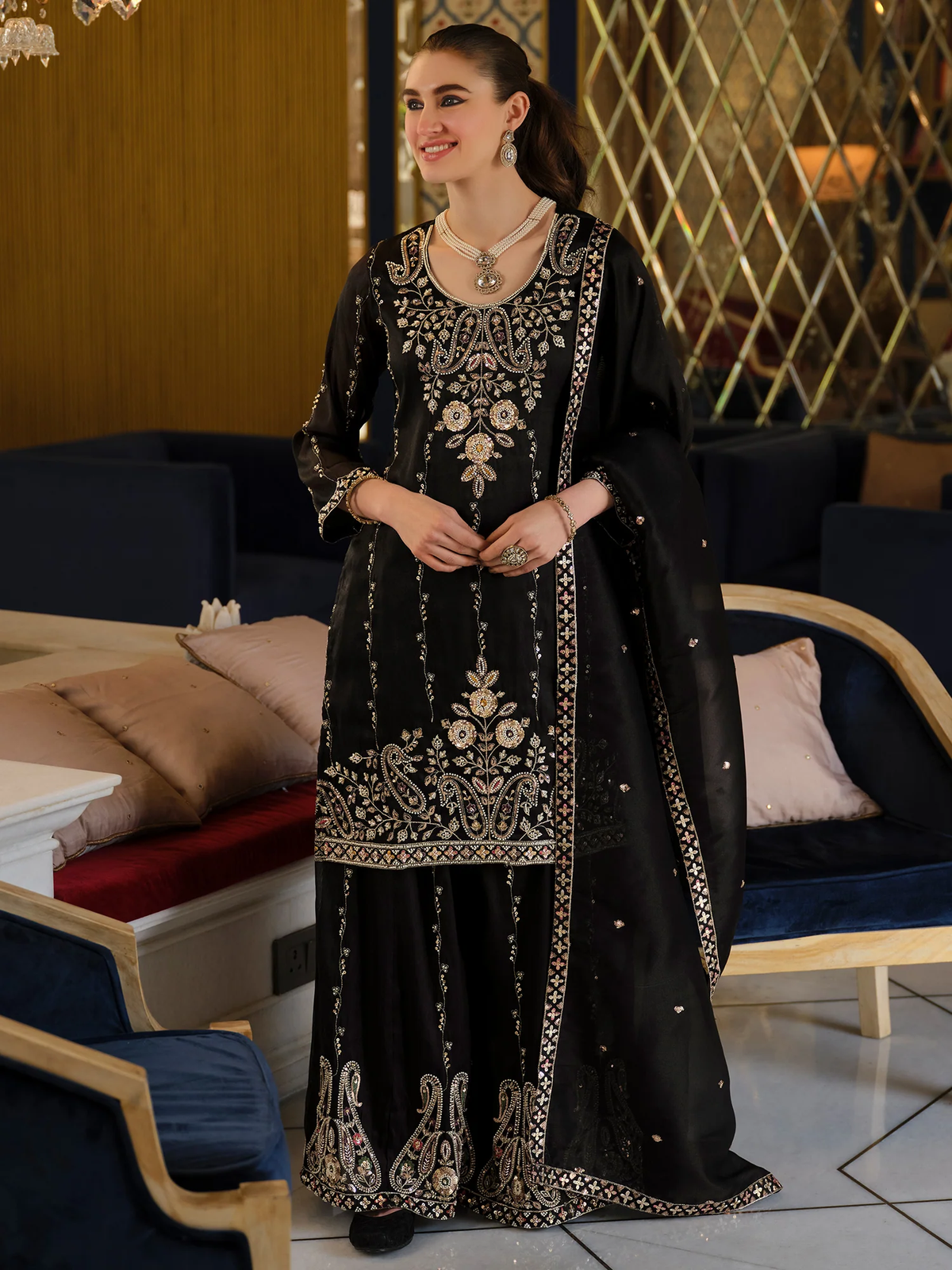 A woman models a black traditional South Asian outfit with extensive silver and gold embroidery, paired with a matching sheer dupatta, in an indoor lounge setting.