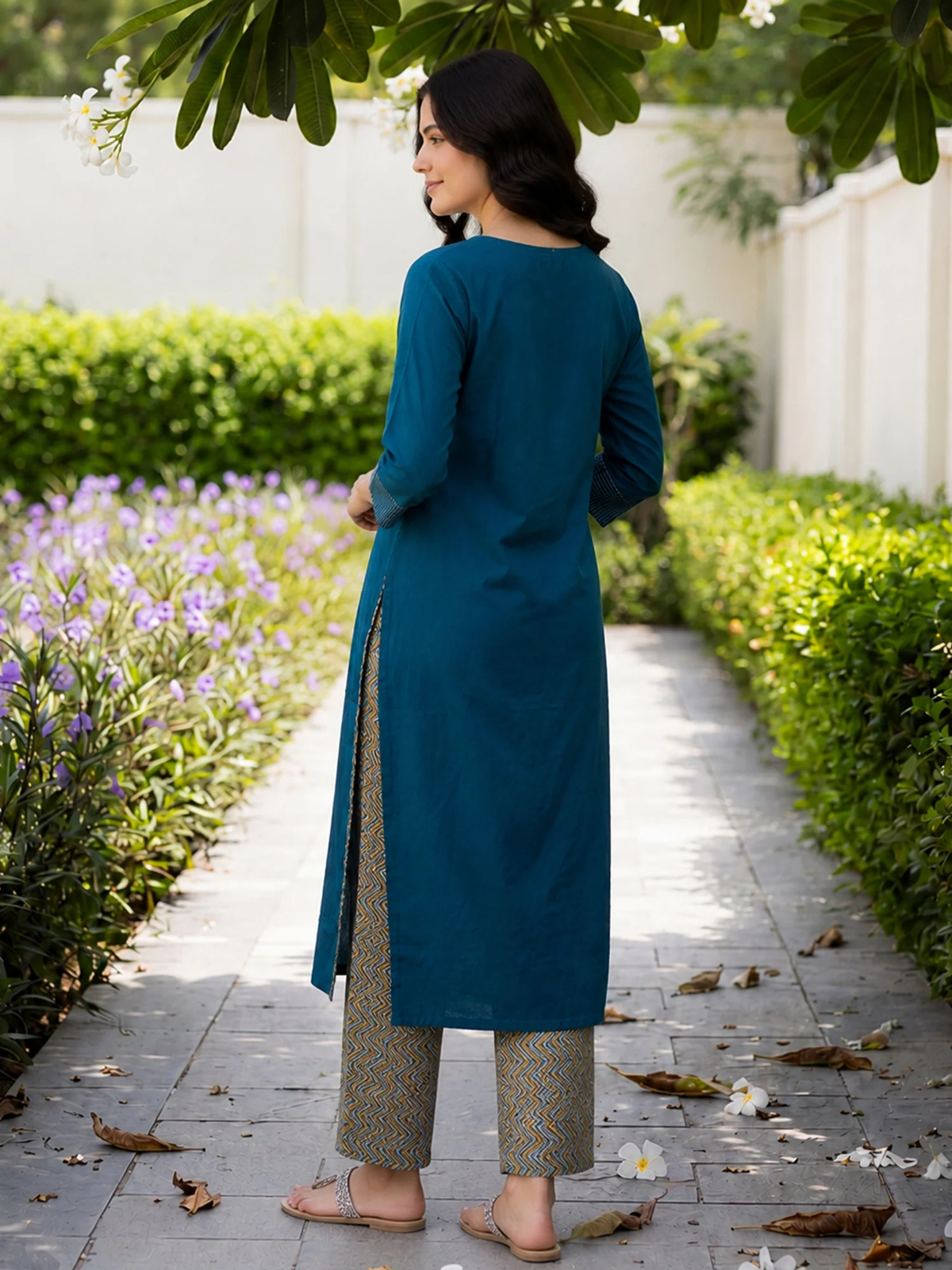 Woman in a teal kurta standing on a garden path with greenery and flowers.