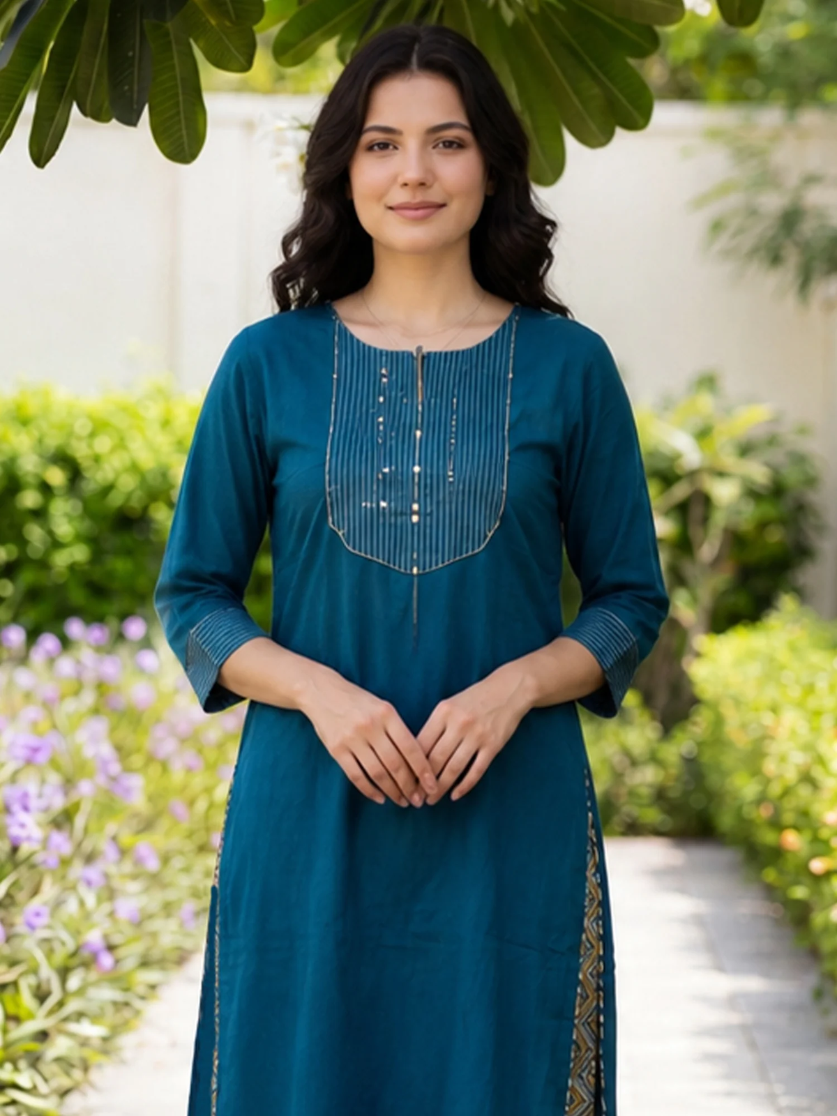 A woman wearing a teal blue kurta with silver beadwork on the chest, standing in a garden setting with green foliage and a light-colored wall in the background.