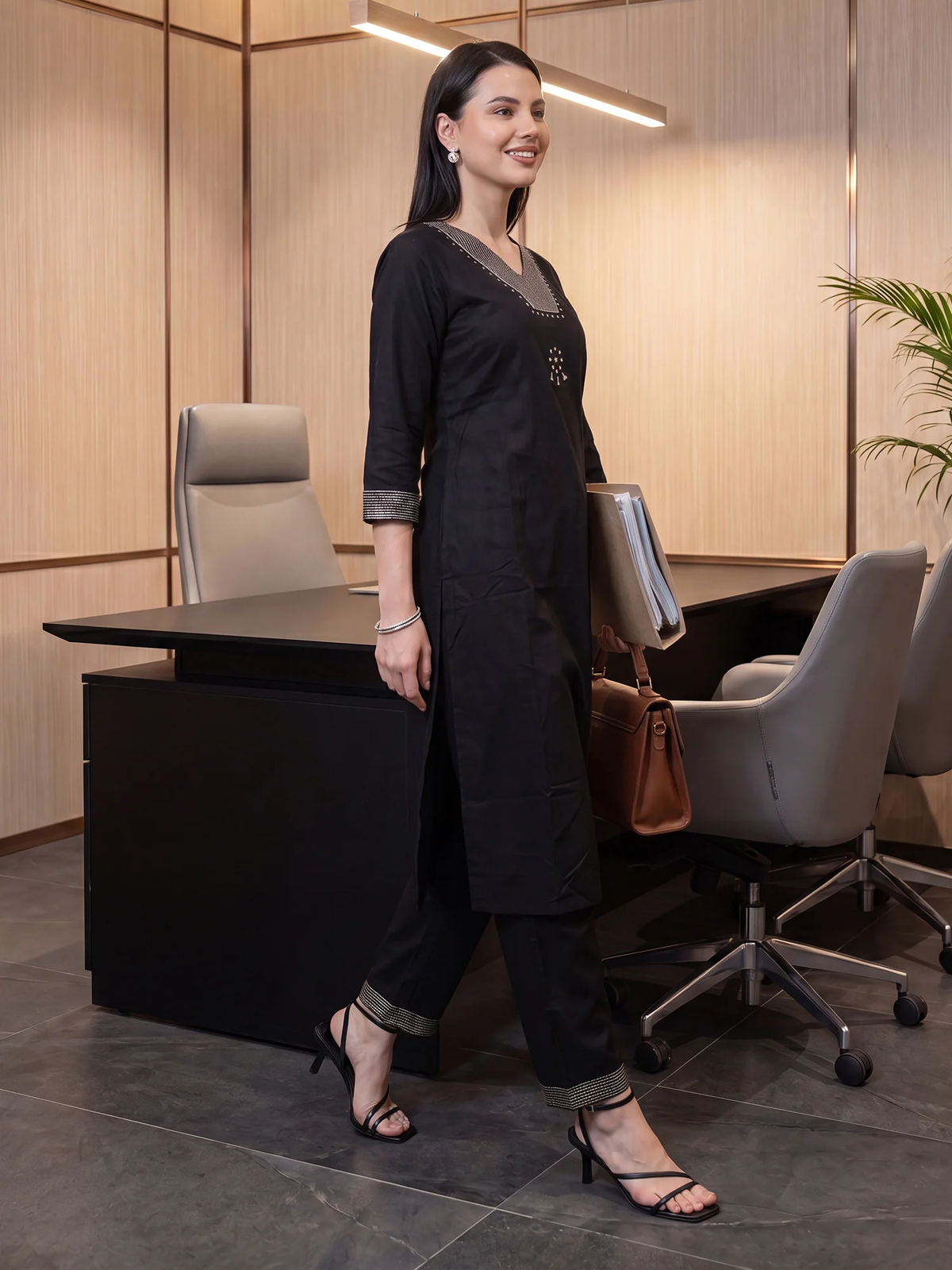 A woman modeling a black three-piece kurta set with matching trousers in a modern office setting. The set features a straight black kurta with a small embroidered detail on the chest, black wide-leg trousers with rolled cuffs, and black heeled sandals.