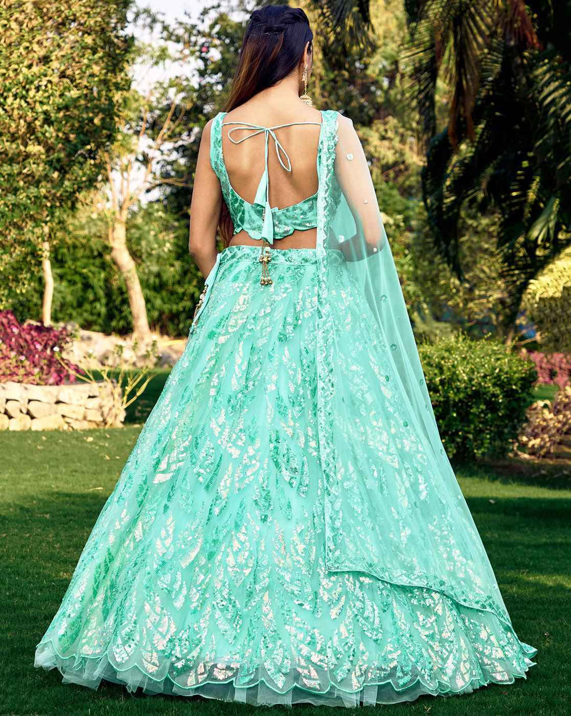 Back view of model in mint green women embellished lehenga choli set with open back tie blouse and sheer dupatta in garden
