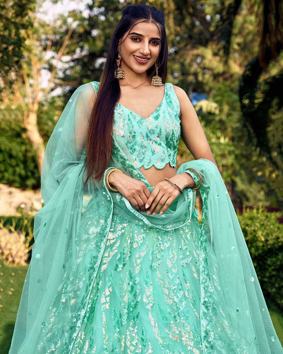 Smiling model in mint green women embellished lehenga choli set with sheer dupatta and statement jhumka earrings in garden