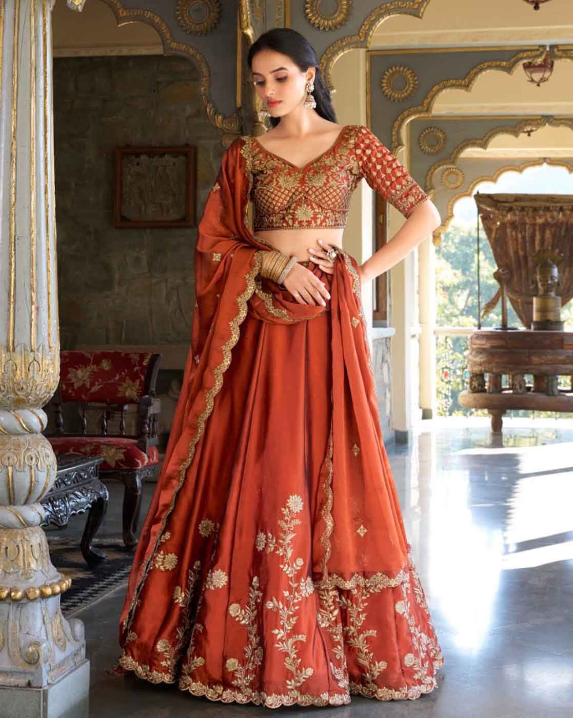 Model wearing rust orange embroidered lehenga choli set Canada with gold floral embroidery and matching dupatta in palace interior