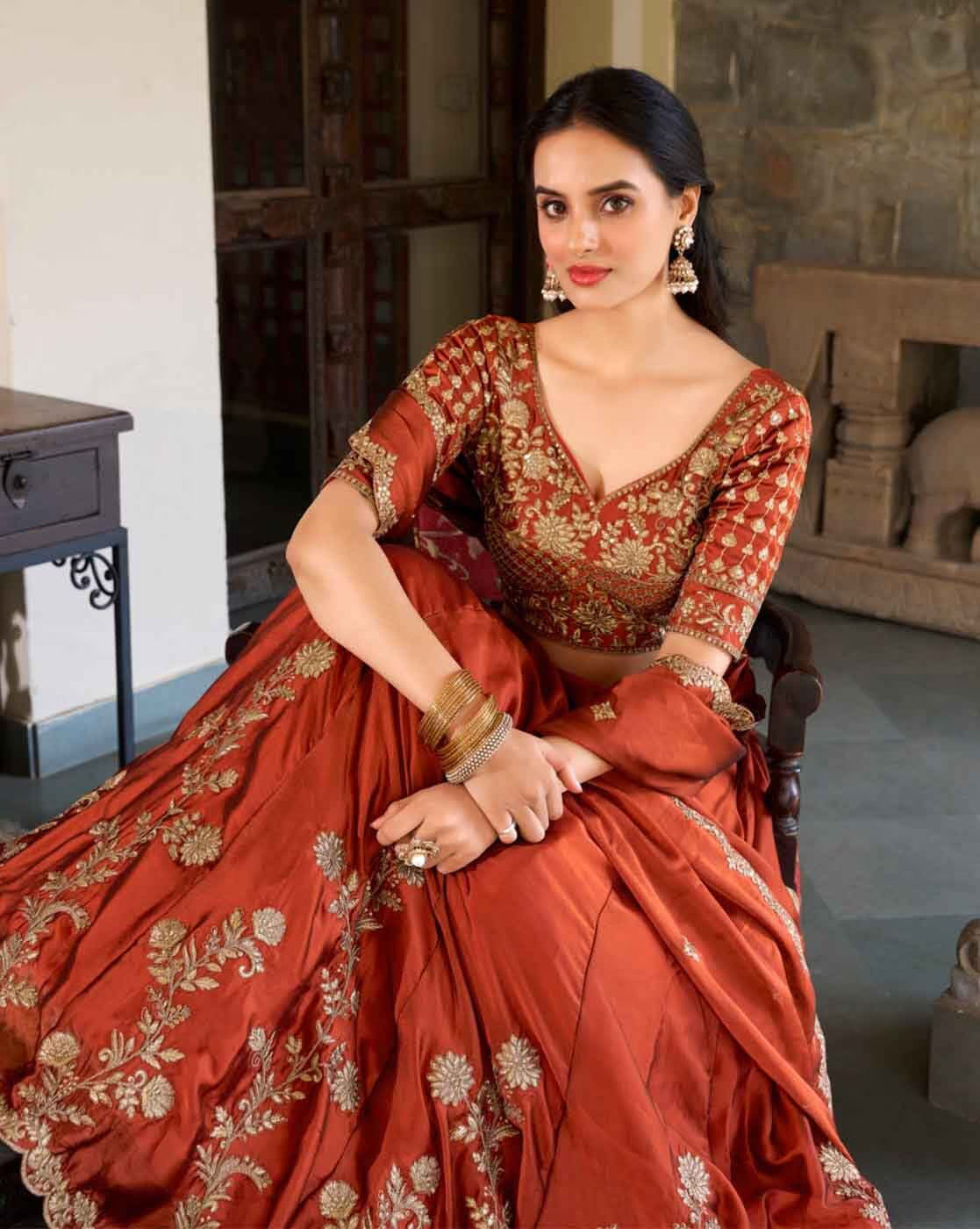 Woman seated wearing rust and gold embroidered lehenga choli set Canada with floral motifs and jhumka earrings