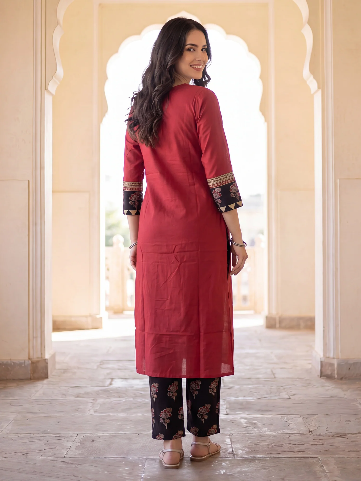 A woman wearing a red long kurta with patterned cuffs and black floral pants, standing in a corridor with arches.