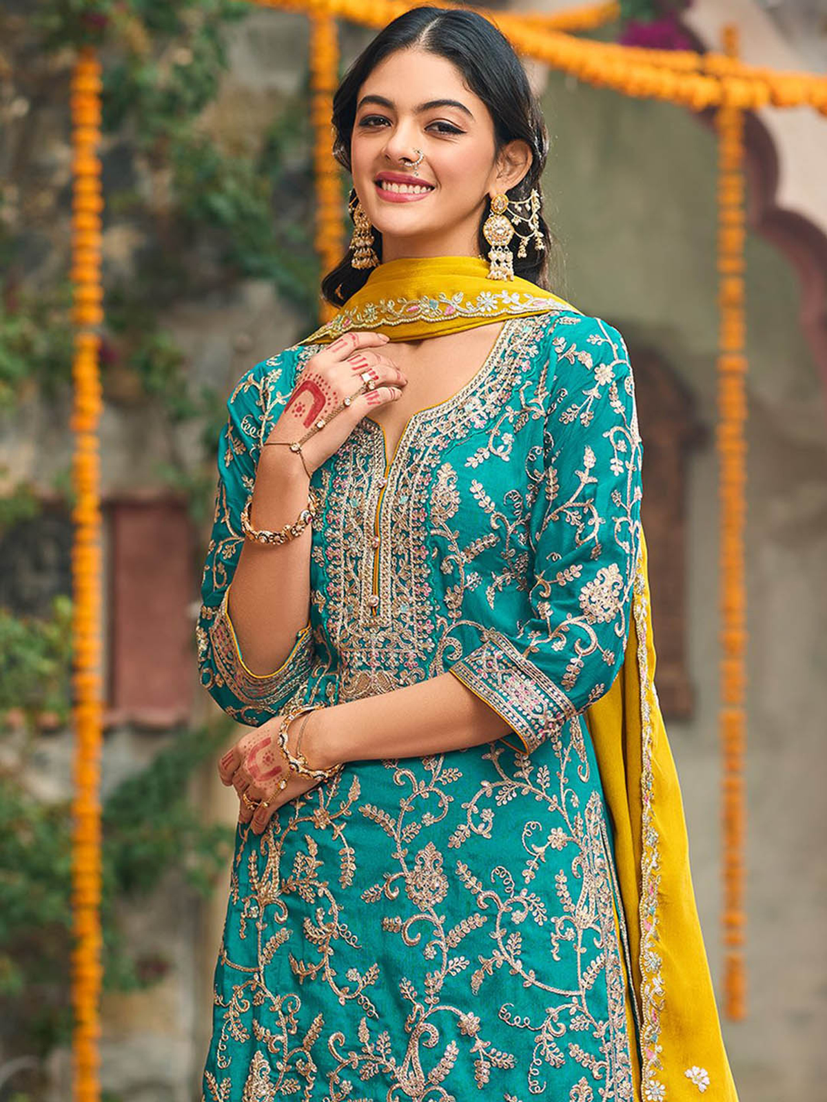 Woman in a teal and yellow traditional outfit with intricate patterns, smiling outdoors.