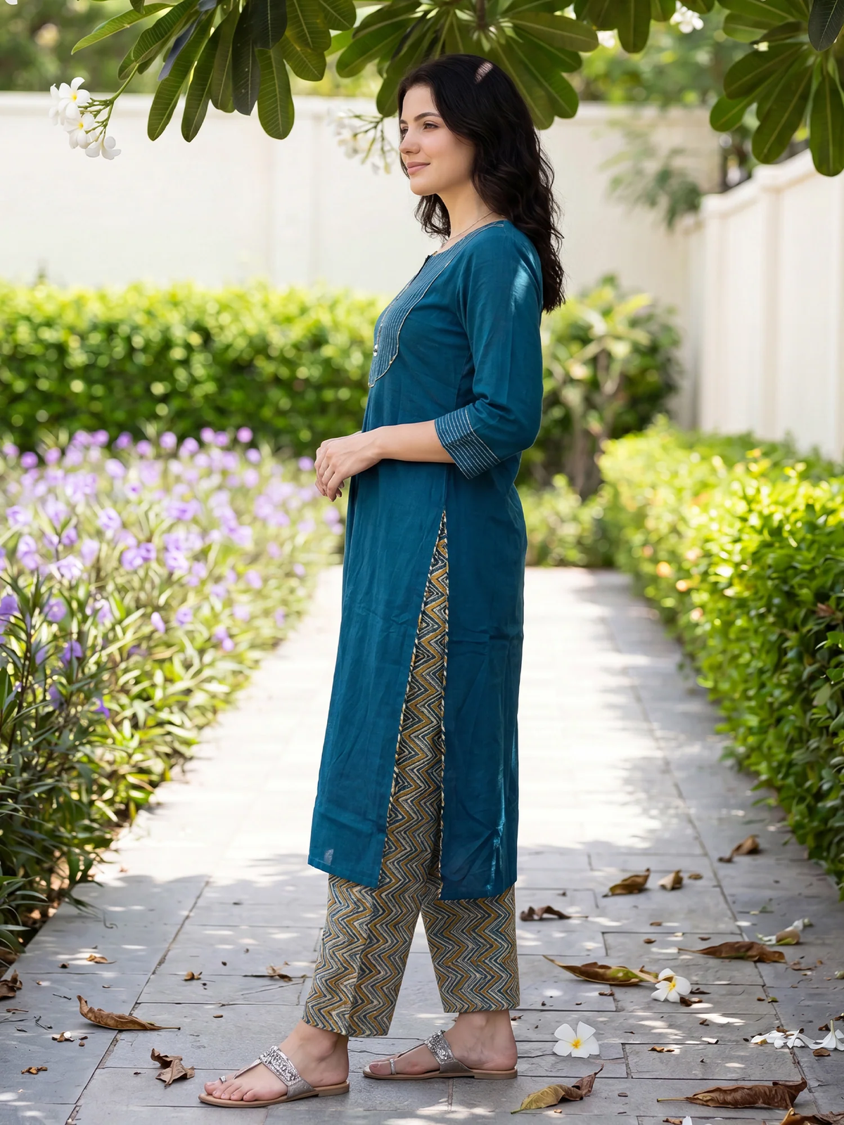 Woman in a teal outfit standing on a garden path with greenery and flowers.