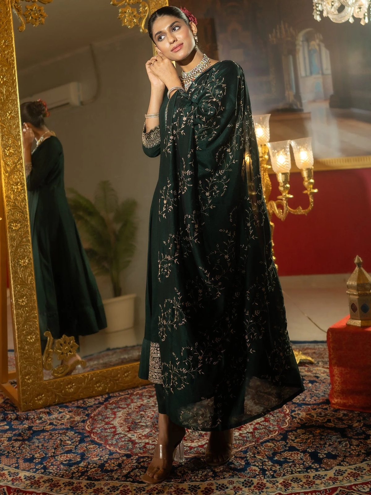 A woman models a dark green traditional outfit with intricate silver floral embroidery, standing in an ornate room with a large gold mirror, patterned rug, and warm lighting.