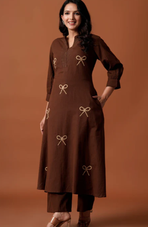 A woman modeling a brown long kurta with matching pants, decorated with beige embroidered bow motifs, set against a solid warm brown studio background.