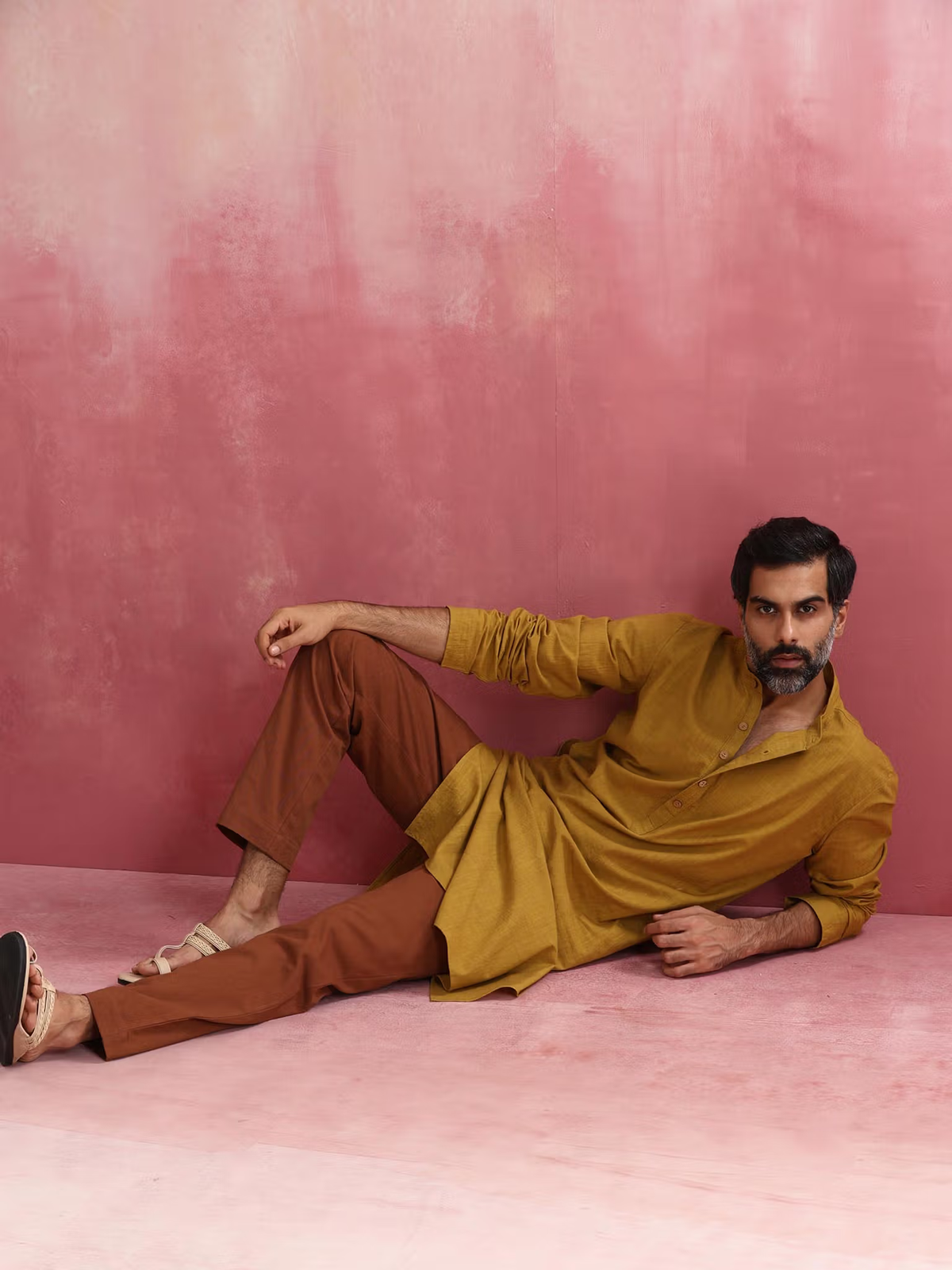 Male model reclining on pink backdrop wearing men's mustard kurta Canada with brown trousers and sandals