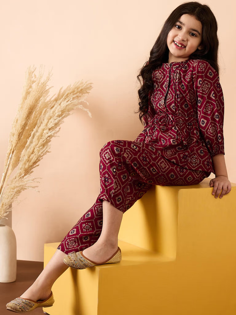 Smiling girl seated on yellow block wearing a maroon printed Girls Georgette Kurta Set with juttis and pampas grass