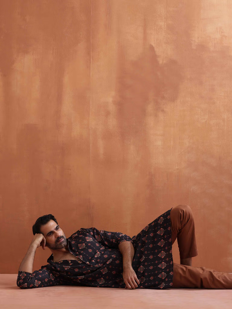 Male model reclining in men's black printed kurta with brown pants on warm studio backdrop