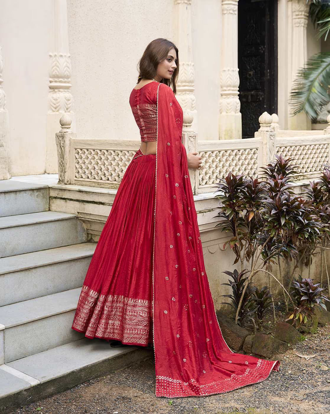 Model wearing red embroidered lehenga choli set with long dupatta trailing on palace steps