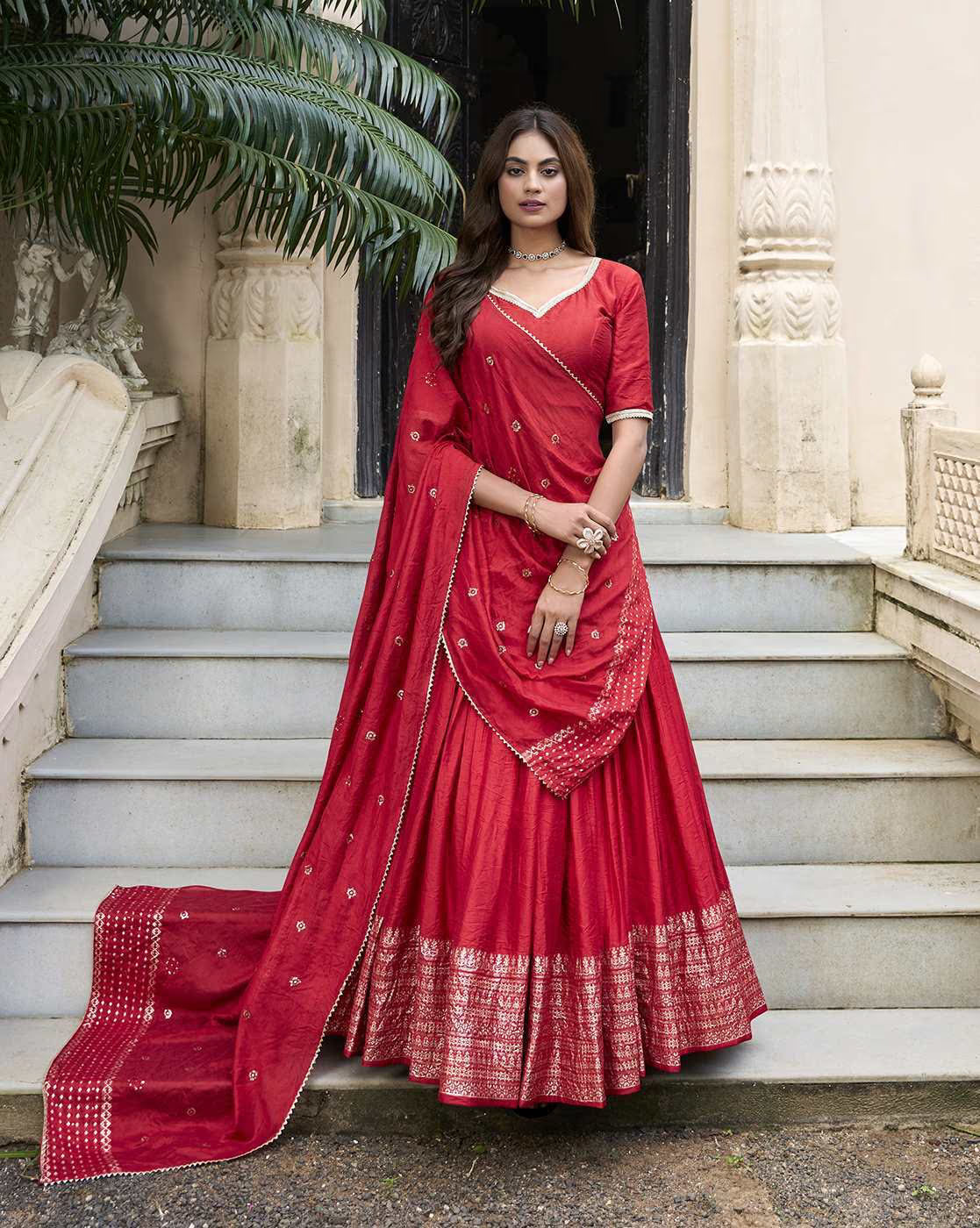 Woman wearing red embroidered lehenga choli set with silver border and dupatta standing on stone steps at historic doorway