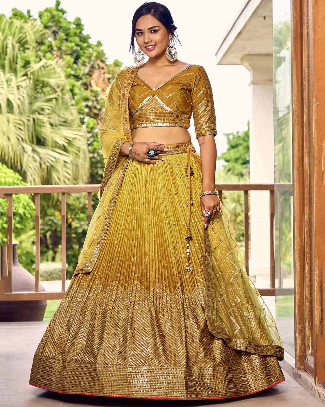 Woman wearing embellished flared lehenga choli Canada in gold with matching dupatta and statement earrings on balcony