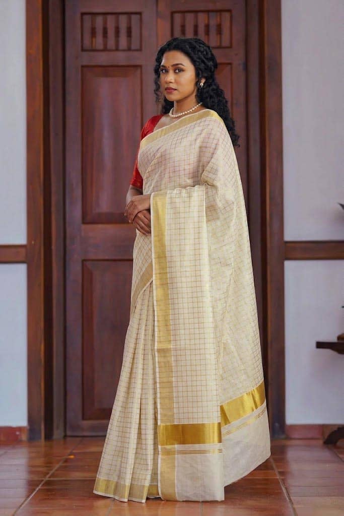 A woman wearing a cream-colored saree with a subtle grid pattern and a gold border, paired with a red blouse, standing in front of a dark wooden door.